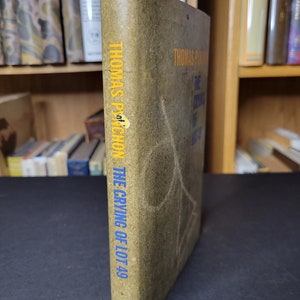 May include: A vintage copy of Thomas Pynchon's "The Crying of Lot 49" with a worn, olive-green cover. The title and author's name are in yellow and blue lettering. The book is displayed with the spine facing the viewer.