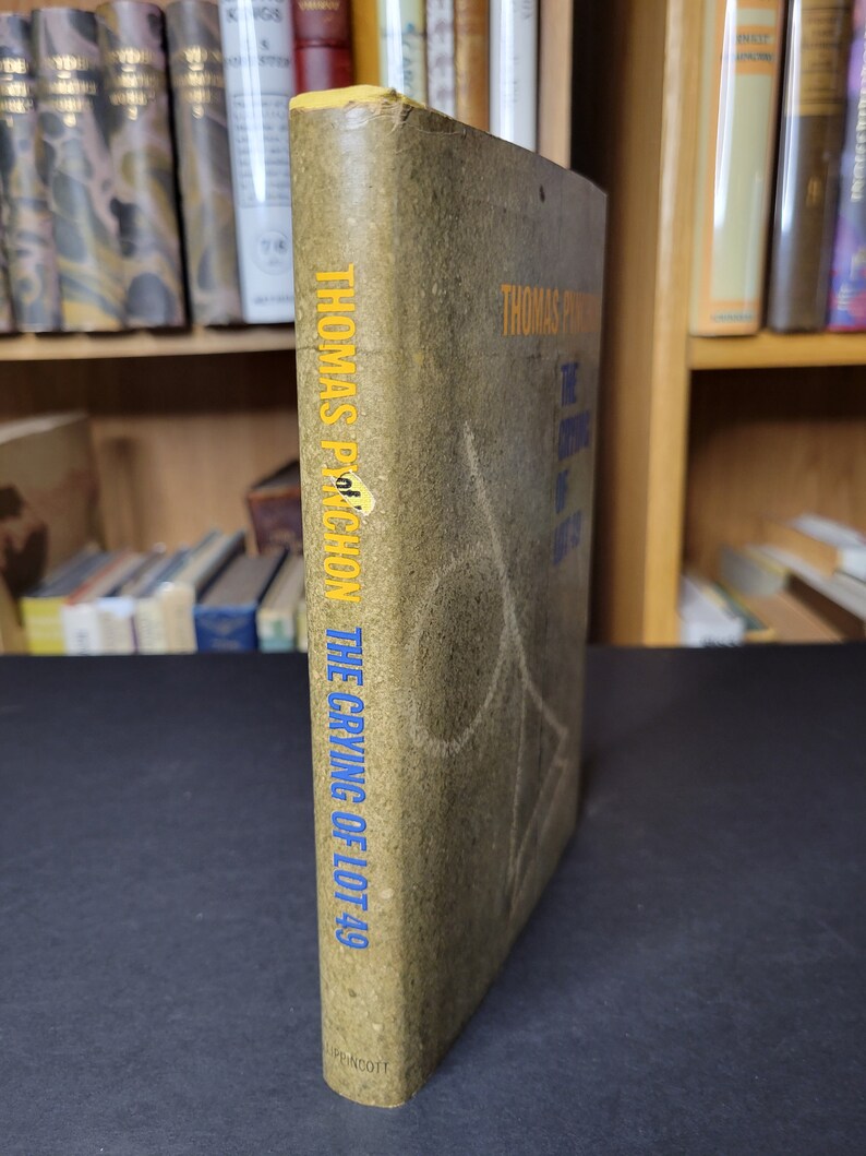 May include: A vintage copy of Thomas Pynchon's "The Crying of Lot 49" with a worn, olive-green cover. The title and author's name are in yellow and blue lettering. The book is displayed with the spine facing the viewer.