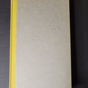 May include: A hardcover book with a light gray cover and a yellow spine. The cover has a textured pattern and embossed trumpet-like designs. The book is closed and lies on a dark surface.