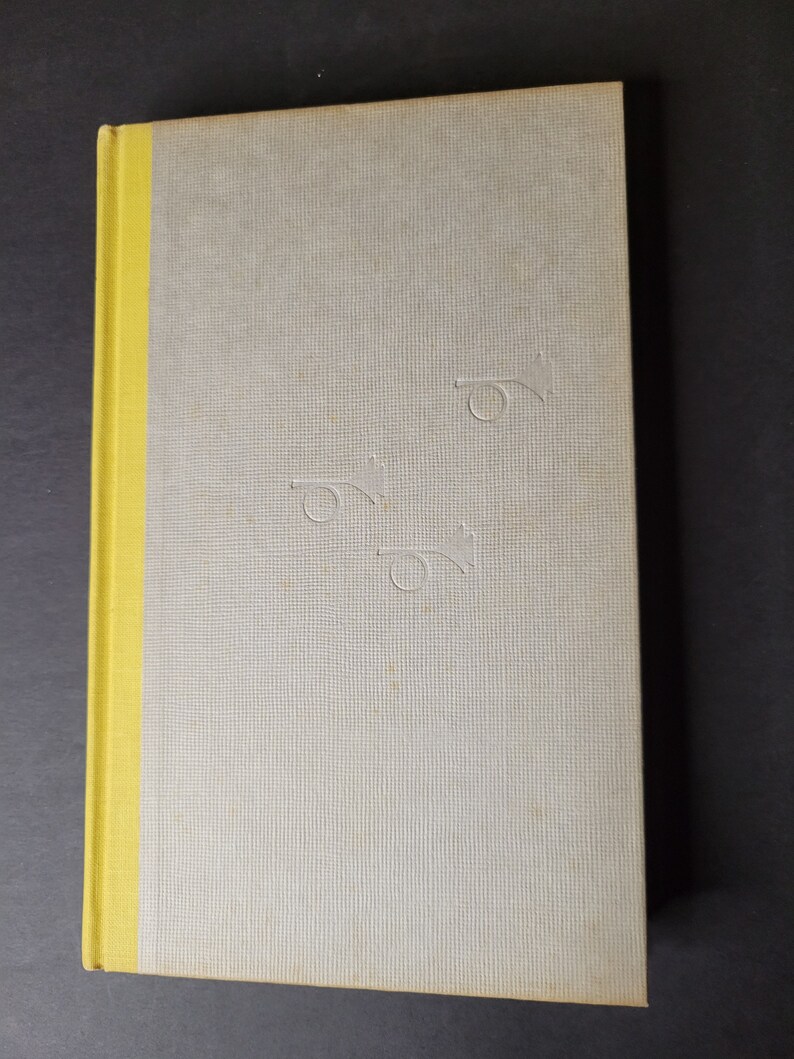 May include: A hardcover book with a light gray cover and a yellow spine. The cover has a textured pattern and embossed trumpet-like designs. The book is closed and lies on a dark surface.