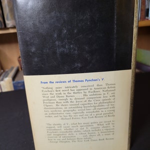 May include: The back cover of a vintage book, featuring a black background and white text. The text includes reviews of Thomas Pynchon's novel "V." from the New York Review of Books and The New York Times Book Review.
