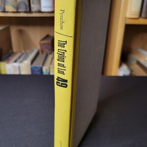 May include: A hardcover book titled "The Crying of Lot 49" by Pynchon. The spine is yellow with black text, and the book's cover is a muted brown. The book is displayed vertically, with the spine facing the viewer. The publisher's name is visible at the bottom of the spine.