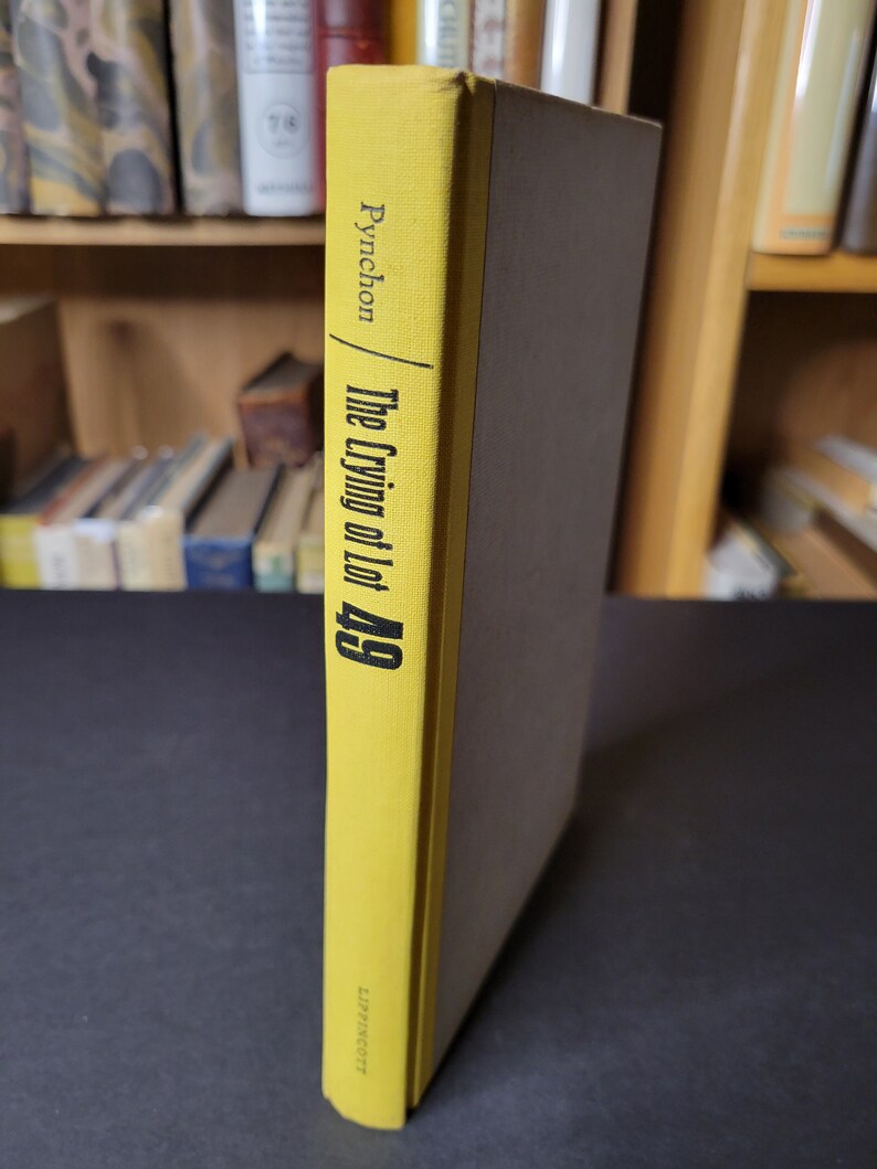 May include: A hardcover book titled "The Crying of Lot 49" by Pynchon. The spine is yellow with black text, and the book's cover is a muted brown. The book is displayed vertically, with the spine facing the viewer. The publisher's name is visible at the bottom of the spine.