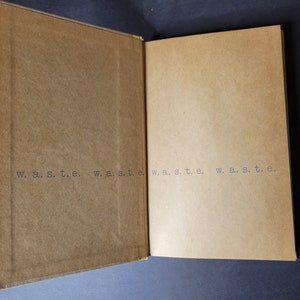 May include: Open book with tan-colored pages and the word "w. a. s. t. e." printed repeatedly in a dark gray font. The book's cover is a darker shade of tan, and the pages are slightly lighter.