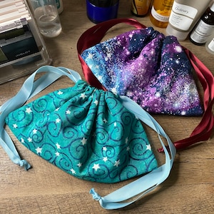 May include: Two fabric drawstring bags with different patterns. One bag is teal with white stars and swirls, and the other is purple with a galaxy print. Both bags have matching ribbon ties.