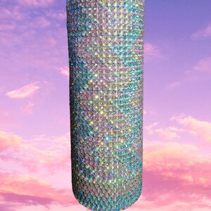 May include: A cylindrical tumbler covered in iridescent rhinestones. The rhinestones are arranged closely together, creating a shimmering effect. The colors shift from clear to shades of blue, pink, and green. The tumbler is set against a pastel sky background.