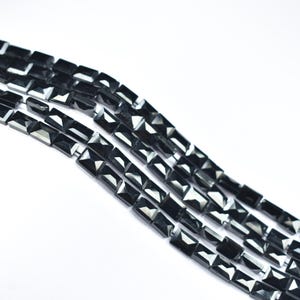 May include: Close-up of several strands of black rectangular beads. The beads have a faceted surface, reflecting light. The beads are strung together, creating a linear pattern against a white background. Ideal for jewelry making.