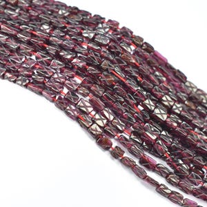May include: Strands of dark red garnet beads with a rectangular shape and a crisscross pattern. The beads are strung on red thread, creating a rich, textured appearance. These beads are suitable for jewelry making.