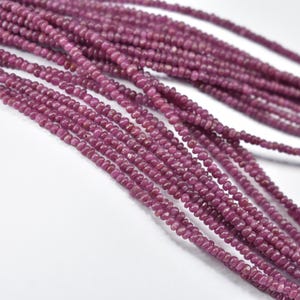 May include: Close-up of multiple strands of small, round, deep pink ruby beads. The beads are strung together, creating a textured, layered effect. The background is a plain white surface, highlighting the vibrant colour of the gemstones.