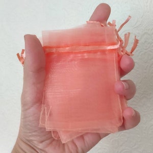 May include: A set of small, peach-colored organza bags with a satin ribbon drawstring. The bags are folded and held in a hand.