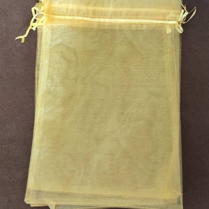 Puede incluir: Varios bolsos transparentes de color amarillo pálido con cordón se apilan sobre una superficie marrón. Las bolsas están hechas de una tela translúcida y los cordones están atados. Las bolsas están vacías.