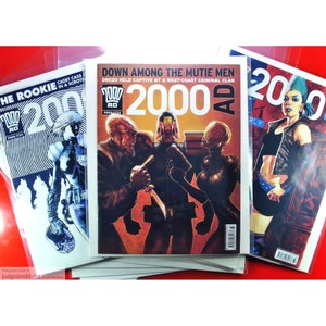 Może przedstawiać: Wyświetlane są trzy komiksy "2000 AD". Centralny komiks zawiera tytuł "Down Among The Mutie Men" i futurystyczną ilustrację. Pozostałe dwa komiksy przedstawiają ilustracje postaci w akcji, z napisami "The Rookie" i "Cadet Cass."