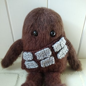 Op de afbeelding: Een bruin, pluche, gehaakt knuffeldier met zwarte ogen en een grijze, gehaakte sjaal. Het speelgoed zit op een wit oppervlak.