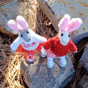May include: Two knitted bunny plushies wearing sweaters and shoes. One bunny is wearing a white sweater with the word "Love" embroidered on it and a red skirt. The other bunny is wearing a red sweater and a plaid skirt.