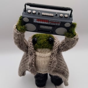May include: A knitted frog toy in a grey coat, cream sweater, and black trousers, holding a black boombox. The frog has a green head and black eyes, with white trainers. The boombox has silver buttons and a red display.