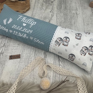 Könnte beinhalten: Ein blaues Kissen mit Waffelstruktur und weißen Stickereien. Das Kissen ist mit dem Namen "Phillip", Fußabdrücken und Geburtsdaten personalisiert. Das andere Ende zeigt einen weißen Stoff mit Pinguin-Illustrationen und einer Spitzenborte.