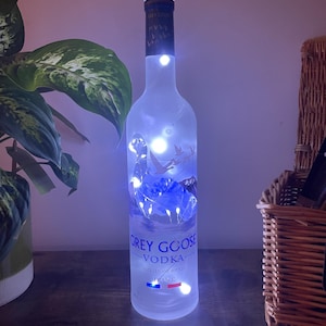 Op de afbeelding: Een hergebruikte Grey Goose wodkafles verlicht met blauwe lichtsnoeren. De fles heeft het merklogo en tekst, met een kurk. De lichten creëren een zachte gloed, waardoor het matte uiterlijk van de fles wordt benadrukt. Een decoratief item.