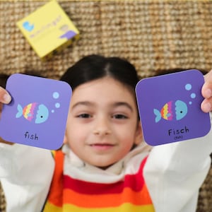 Pu&ograve; includere: Due carte educative quadrate viola raffiguranti un pesce dei cartoni animati con il corpo color arcobaleno e la testa blu. Una carta riporta la parola fish, l'altra la parola Fisch con la traduzione inglese tra parentesi sotto.