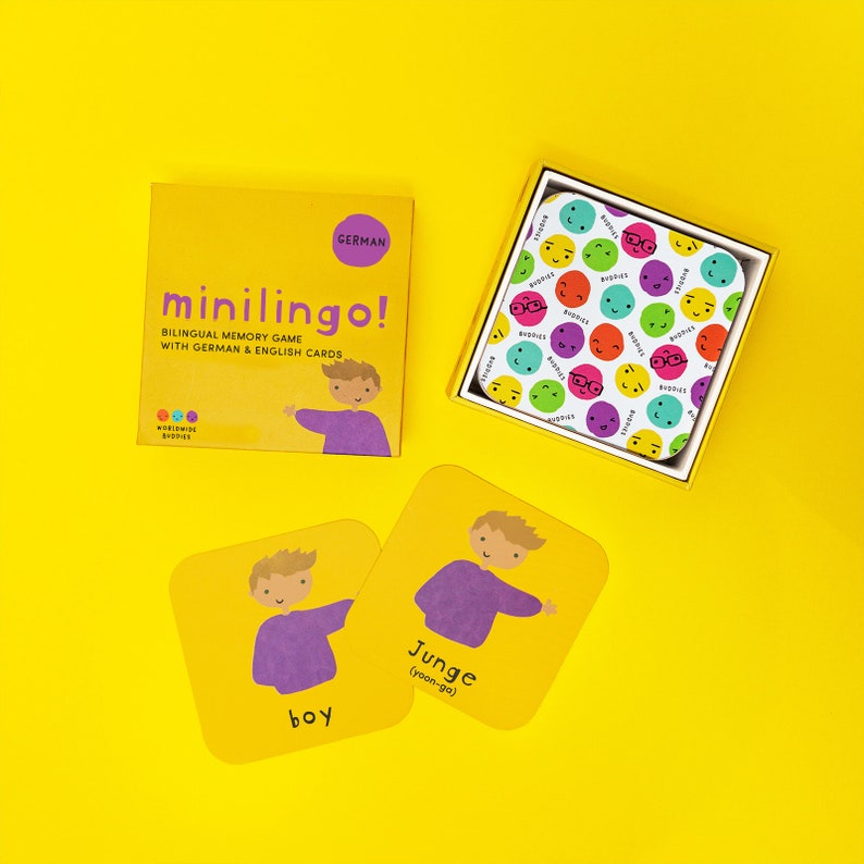 Pu&ograve; includere: Un gioco di memoria bilingue Minilingo! con una scatola gialla che mostra un personaggio, il testo "German" e "Bilingual memory game with German & English cards." Include una scatola di carte con motivi di facce colorate e due carte gialle che mostrano un ragazzo.