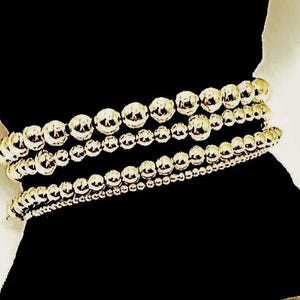 May include: A set of four stacked bracelets featuring polished gold-tone spherical beads of varying sizes. The bracelets are arranged in a row, showcasing a reflective metallic finish on each round bead, creating a uniform and consistent appearance.