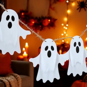 May include: A Halloween-themed garland of three white paper ghost cutouts with black eyes and mouths, strung on twine. The background features warm lighting, a decorative spider, and pumpkins, creating a festive atmosphere.