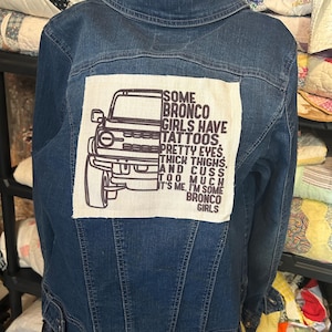 May include: A dark blue denim jacket with a patch on the back. The patch features a line drawing of a Bronco SUV and the text: "SOME BRONCO GIRLS HAVE TATTOOS. PRETTY EYES. THICK THIGHS. AND CUSS TOO MUCH. IT'S ME. I'M SOME BRONCO GIRLS."