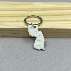May include: White ceramic keychain shaped like the state of New Jersey with a silver key ring. The keychain has the word "NJ" embossed on it.