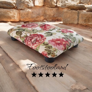 May include: A floral patterned footstool with a cream background and red, pink, and green flowers. The footstool has four black wooden legs.