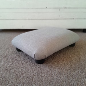 May include: A light grey fabric footstool with four black legs. The footstool is sitting on a light brown carpet.