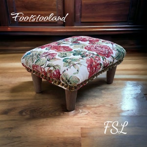 May include: A floral patterned footstool with a wooden frame and four legs. The fabric features a white background with red and green floral designs. The footstool is sitting on a wooden floor.