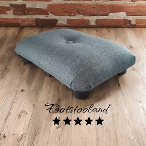 Op de afbeelding: Een grijze stoffen voetenbank met een knoop in de bekleding bovenop en vier zwarte houten poten. De voetenbank staat op een houten vloer. De tekst "Footstooland" en vijf sterren zijn zichtbaar onder de voetenbank.