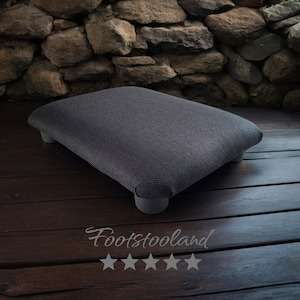 May include: A dark gray rectangular footstool with a textured fabric cover and four wooden legs. The footstool is sitting on a wooden floor.