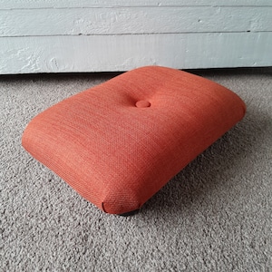May include: A rectangular orange footstool with a button tuft on top. The footstool is made of a textured fabric and has a dark wood base.