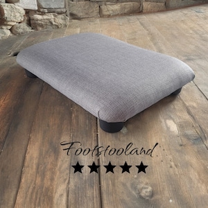 May include: A gray fabric footstool with black wooden legs. The footstool is sitting on a wooden floor. The text "Footstooland" is visible below the footstool.
