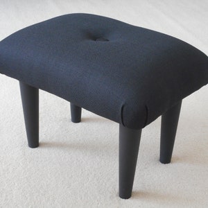 May include: A black upholstered footstool with four short, black legs. The stool has a button tuft on the top.