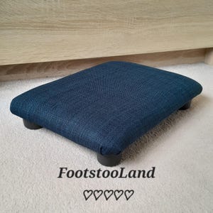 May include: A navy blue footstool with a rectangular shape and four black legs. The footstool is upholstered in a textured fabric. The text "FootstoolLand" and five hearts are printed on the bottom.