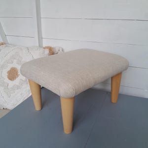 Op de afbeelding: Een kleine, rechthoekige voetenbank met een lichtbeige stoffen zitting en vier houten poten. De poten hebben een lichte, natuurlijke houtkleur. De voetenbank staat op een blauwgrijs oppervlak.