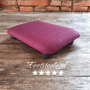 May include: A small, rectangular footstool with a dark purple fabric cover and black wooden legs. The footstool is sitting on a wooden floor.