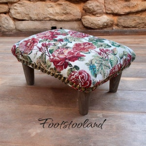 Puede incluir: Un reposapiés con estampado floral de fondo blanco y flores rojas, rosas y verdes. El reposapiés tiene cuatro patas de madera y un ribete dorado alrededor del borde superior.