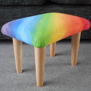 May include: A rainbow-coloured footstool with four wooden legs. The footstool is made of fabric and has a gradient design that starts with purple and ends with red.
