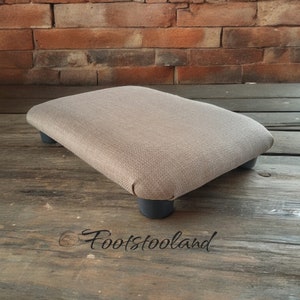 May include: A light brown fabric footstool with four black legs. The footstool is sitting on a wooden surface.