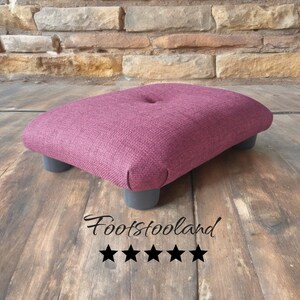 May include: A burgundy fabric footstool with four dark grey legs. The footstool has a button tuft on the top. The text "Footstooland" and five stars are visible below the footstool.