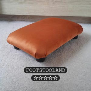 Könnte beinhalten: Ein orangefarbener Samt-Fußhocker mit schwarzen Beinen. Der rechteckige Hocker hat abgerundete Kanten und steht auf einem hellgrauen Teppich. Der Text "FOOTSTOOLAND" wird darunter angezeigt.