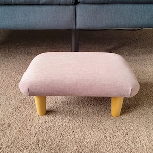 May include: A light pink upholstered footstool with wooden legs. The footstool is sitting on a beige carpet.