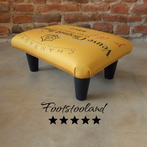 May include: A yellow footstool with black legs. The footstool is covered in a fabric with the Veuve Clicquot Champagne label. The label reads "Veuve Clicquot Ponsardin, Reims, France, Brut, Appreciation & Originale, Champagne."