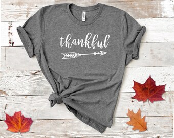 Puede incluir: Una camiseta gris con la palabra "thankful" escrita en letras blancas cursivas con una flecha blanca apuntando hacia la derecha. La camiseta está anudada y se muestra sobre un fondo de madera blanca con hojas de otoño.