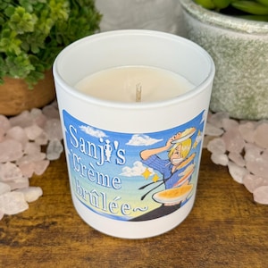 Könnte beinhalten: Weiße Kerze in einem Glasbehälter mit dem Text "Sanji's Crème Brûlée" und einer Illustration einer Figur. Die Kerze ist von dekorativen Elementen umgeben, darunter Pflanzen und rosa Kristalle.