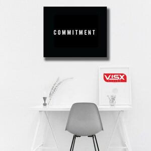 May include: A minimalist office setup featuring a white desk, a gray chair, and two framed artworks. The top artwork is a black square with the word "COMMITMENT" in white. The second artwork has a red logo.