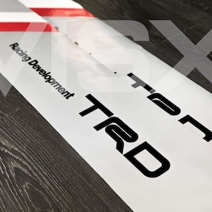 Może przedstawiać: Biała naklejka z czarnym tekstem, który brzmi "TRD" i "Racing Development".