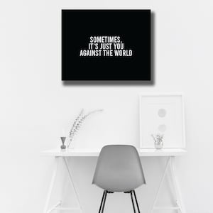 May include: A black framed wall art with the text "SOMETIMES, IT'S JUST YOU AGAINST THE WORLD" in white. The art hangs above a white desk with a gray chair. A framed print and decorative items are on the desk.